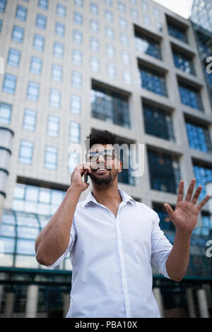 Giovane bello Afro American uomo in piedi di fronte a un enorme e moderno edificio aziendale; sorridente e parlando al telefono cellulare. Foto Stock