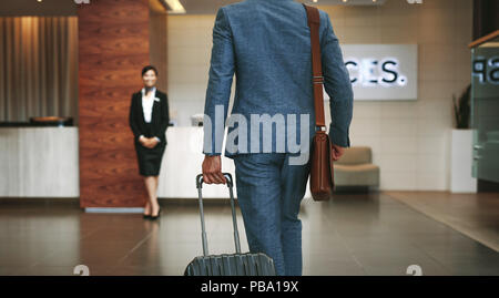 Imprenditore valigetta mentre si cammina nella lobby dell'hotel. Per chi viaggia per affari che arrivano in hotel con addetta alla ricezione in piedi in background per noi Foto Stock