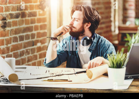 Concentrato giovane architetto lookingto la finestra e pensare a nuovi blueprint durante la seduta con il computer portatile in un loft sole ufficio Foto Stock