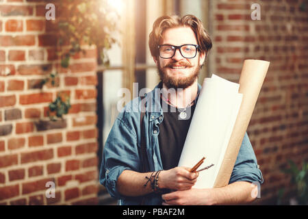 Attractiwe barbuto dark-capelli architetto maschio che indossano occhiali azienda blueprint e sorridenti vicino alla finestra in mansarda ufficio moderno Foto Stock