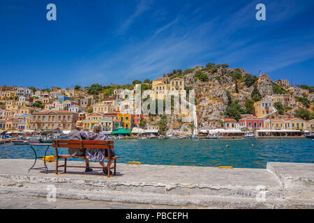 Scenic vista romantica con una vecchia coppia seduta su una panchina guardando verso le belle case tradizionali e gli edifici colorati al porto di Symi Foto Stock