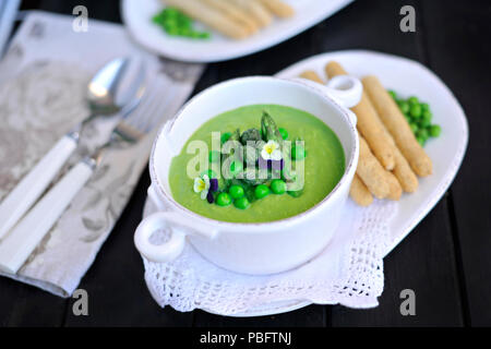 Zuppa con purea di piselli verdi e asparagi. decorate con fiori freschi violette. su uno sfondo nero. in bianco piatti. pane e fette biscottate in un serving. Foto Stock