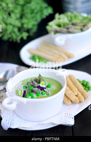 Zuppa con purea di piselli verdi e asparagi. decorate con fiori freschi violette. su uno sfondo nero. in bianco piatti. pane e fette biscottate in un serving Foto Stock