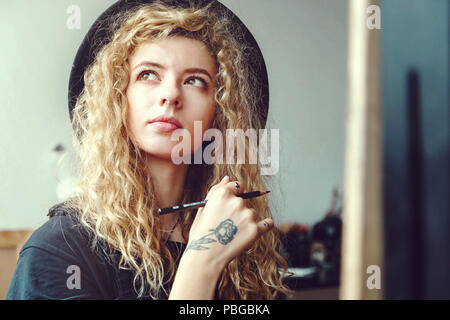 Ritratto di lunghi capelli ricci ragazza nel bel black hat seduto e sognare con matita dietro il cavalletto in studio Foto Stock