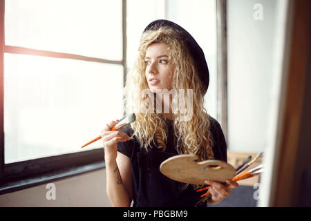 Romantico biondi capelli lunghi artista femminile in bella black hat holding pennello e tavolozza dietro il cavalletto in studio vicino alla finestra Foto Stock