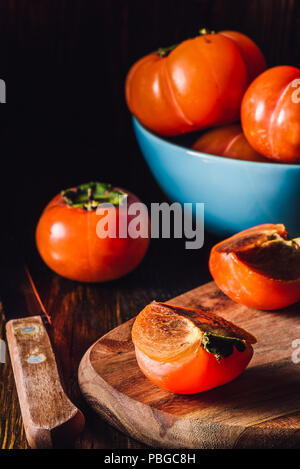 Persimmon fette con il coltello e alcuni frutti in vaso blu su sfondo. Verticale. Foto Stock