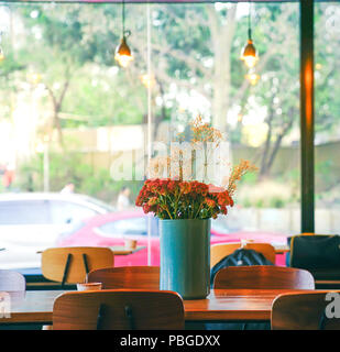 Un mazzetto di fiori di colore rosso decorata in blu vaso in ceramica sul tavolo in legno con sedie in legno, nel mezzo del cafe con due luce arancione lampadine o sospesi Foto Stock