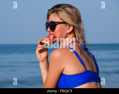 Una bionda in occhiali da sole mangia un cocomero dal mare. Una succosa anguria nelle mani di una donna. Foto Stock