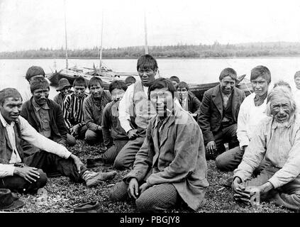 Indian uomini e ragazzi in abito occidentale, seduti lungo il litorale, barche in background. 1900-1923 della Baia di Hudson indiani Foto Stock