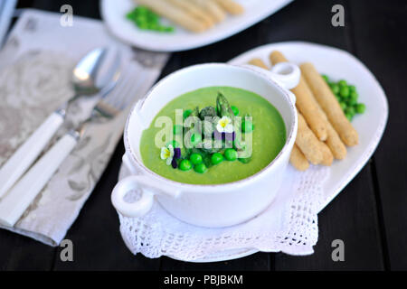 Zuppa con purea di piselli verdi e asparagi. decorate con fiori freschi violette. su uno sfondo nero. in bianco piatti. pane e fette biscottate in un serving Foto Stock