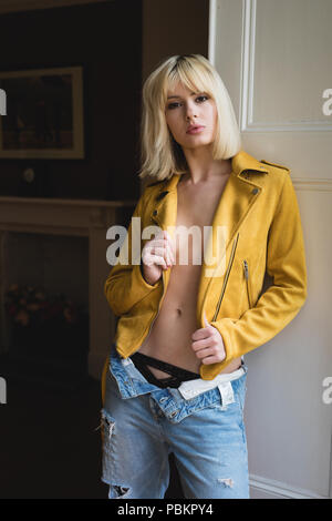 Donna bionda in piedi accanto a una finestra con persiane indossando un giallo senape giacca di pelle. Le sue mani sono tenendo la camicia aperta. Foto Stock