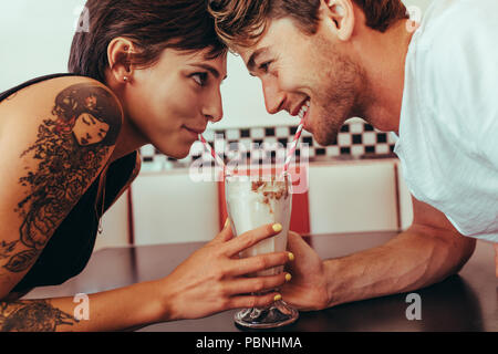 In prossimità di una coppia felice sorseggiando milk shake da un vetro con cannucce mentre guardando ogni altro. L uomo e la donna condividere milk shake utilizzando due cannucce Foto Stock