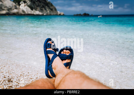 Giovane maschio feets incrociate in blu flip-flop sandalo a prendere il sole sulla spiaggia del mare Foto Stock