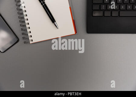 Moderno edificio grigio office desk tabella con lo smartphone, vuoto pagina del notebook con la penna e la tastiera. Lavorando, concetto di scrittura Foto Stock