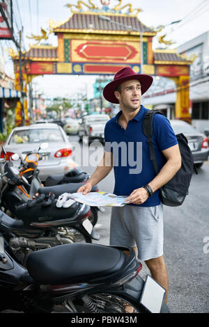 Viaggiatore con una mappa nelle mani sorge sulla strada di città e guarda al lato. Egli indossa una polo blu, grigio pantaloncini, crimson hat e uno zaino nero. Foto Stock