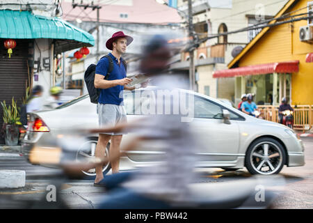 Abbastanza turistico con una mappa nelle mani sorge sulla strada di città e guarda al lato. Egli indossa una polo blu, grigio pantaloncini, crimson hat e un nero bac Foto Stock