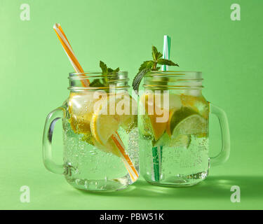 Agrumi fette di limone e lime, ghiaccio, acqua e cannucce di plastica in vetro su uno sfondo verde. Due vasetti di vetro con un freddo naturale limonata fatta a mano con bolle d'aria. Foto Stock