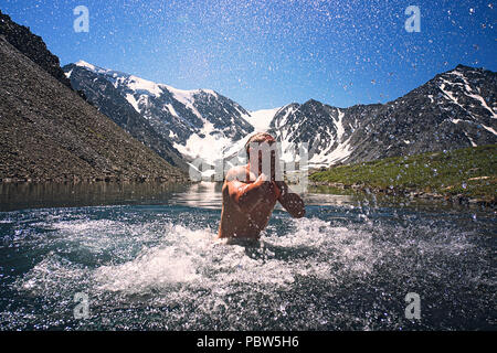 Giovane uomo a nuotare in un lago di montagna sullo sfondo del bellissimo paesaggio di montagna con cime innevate. Intrattenimento estremo. indurire in wate a freddo Foto Stock