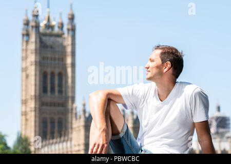 Felice giovane uomo seduto sulla ringhiera a Londra, guardando il cityscape skyline della città con il fiume Tamigi, il Palazzo di Westminster, edificio nel corso estivo soleggiato Foto Stock