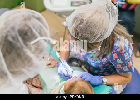 Bambino con una madre a un dentista di ricezione. La ragazza si trova in cattedra, dietro la madre. Il dottore lavora con un assistente. Procedura per la perforazione di un dente. Impostazione di una sigillatura temporanea. Foto Stock