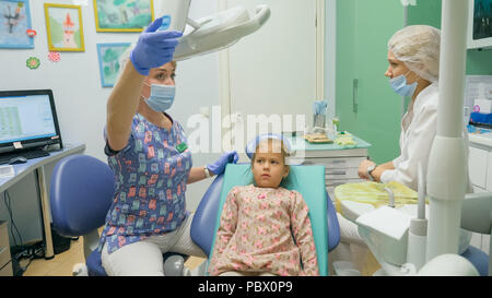 Bambino con una madre a un dentista di ricezione. La ragazza si trova in cattedra, dietro la madre. Il dottore lavora con un assistente. Procedura per la perforazione di un dente. Impostazione di una sigillatura temporanea. Foto Stock