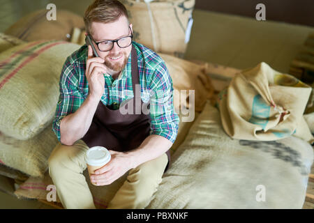 Angolo alto ritratto del bel giovane uomo seduto sul mucchio di sacchi di tela tenendo la carta tazza di caffè e di parlare al telefono tenendo break in modalità Foto Stock