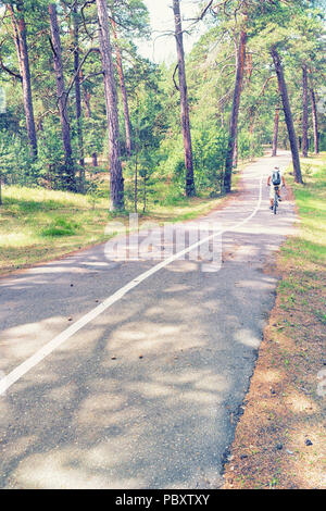 Un ciclista viaggia lungo l'asfalto percorso in bicicletta con una banda bianca in una foresta di pini in una soleggiata giornata estiva Foto Stock