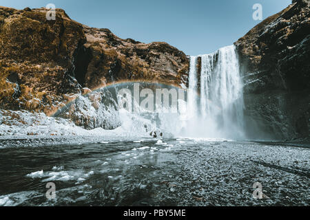 Skogafoss cascata in Islanda con arcobaleno su una giornata soleggiata con cielo blu. Foto Stock