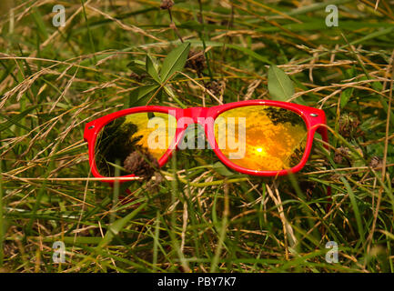 Nuvole e sole riflesso nel vetro di occhiali da sole rosso con telaio in plastica su un prato in erba Foto Stock