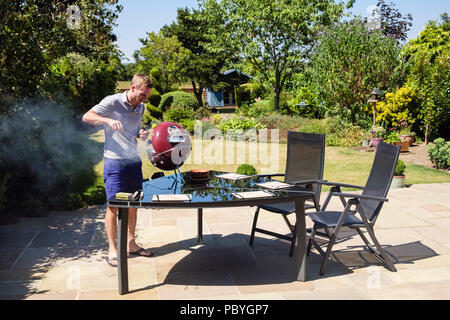 Autentica immagine di un giovane uomo Millenario che cucina su un barbecue su un patio giardino cortile tavolo in una calda stagione estiva del 2018. Inghilterra, Regno Unito, Gran Bretagna Foto Stock