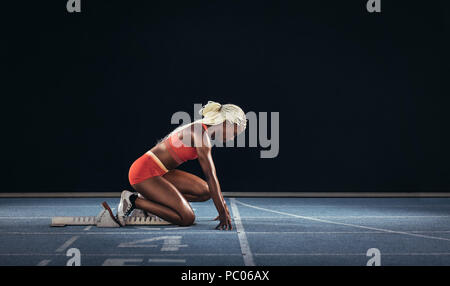 Atleta femminile di prendere posizione sul suo segna per iniziare la corsa. Vista laterale del femminile ottenere pronto all'inizio linea sulla via di corsa su un bl Foto Stock