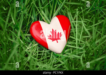Macro closeup colpo di legno cuore di piccole dimensioni con rosso bianco bandiera canadese maple leaf giacente in erba sulla foresta verde natura al di fuori di sfondo, Canada giorno Foto Stock
