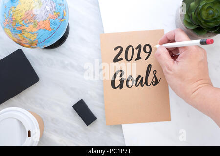 Mano che tiene la penna scrittura 2019 gli obiettivi sulla carta marrone con blue globe,blackboard,tazza di caffè sul tavolo di marmo.risoluzioni del nuovo anno concept Foto Stock