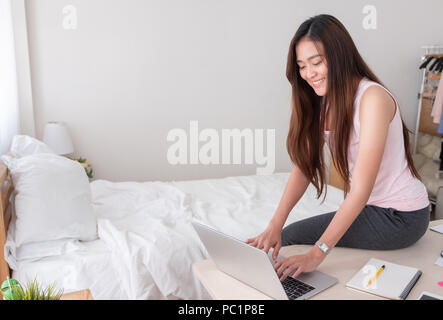 Femmina asiatica del freelancer giubbotto di usura shirt digitazione sul computer portatile sul tavolo e pensare al lavoro nella camera da letto di casa.Il lavoro a casa.facendo scartoffie da casa Foto Stock