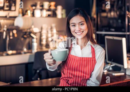 Donna asiatica barista usura grembiule rosso tenendo calda tazza di caffè e sorridente al banco bar con happy emozione,Cafe Restaurant service concetto,focu selettiva Foto Stock