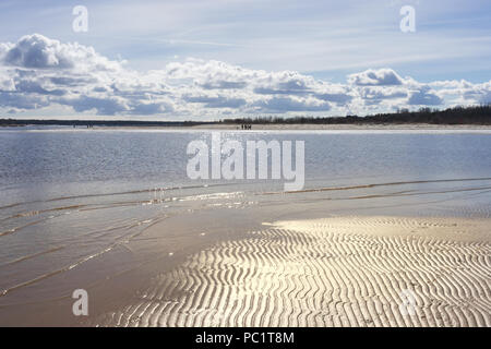 La gente a piedi lungo la riva sabbiosa del fiume con i riflessi dell'acqua su una soleggiata giornata di primavera Foto Stock