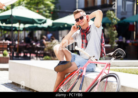 Bel giovane hipster uomo in occhiali da sole seduta vicino alla bicicletta e utilizza lo smartphone nel soleggiato street Foto Stock
