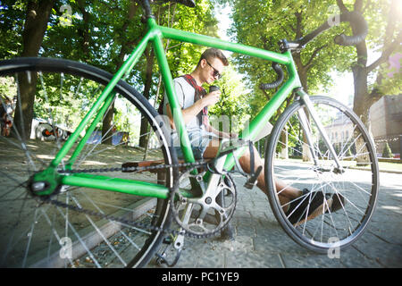 Bello hipster boy in occhiali da sole con bicicletta verde seduto su beanch e lavorare con il computer portatile nella soleggiata estate park, egli a bere caffè Foto Stock