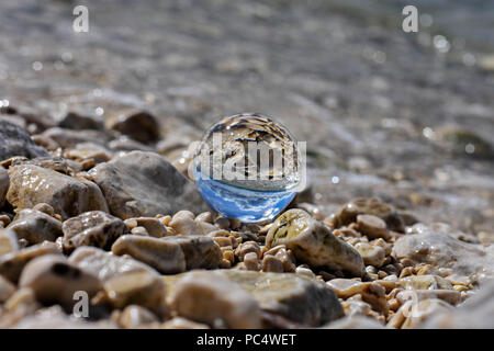 Palla di vetro seduto alla spiaggia rocciosa whit mare onda riflessa in esso/ immagine concettuale delle vacanze estive Foto Stock