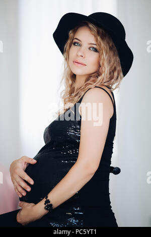 Ritratto in studio di attraente donna bionda con professional make up abbracciando il suo ventre in attesa di baby, indossa una parte superiore nera ed elegante black hat Foto Stock