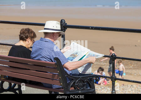 Coppia di anziani seduti sul sedile che si affaccia sulla spiaggia. L'uomo leggendo il quotidiano. Estate 2018. Regno Unito Foto Stock