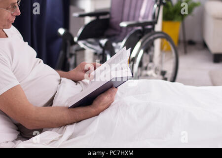 Età compresa tra Uomo con occhiali lettura libro nel letto. Immagine ravvicinata di uomo sfogliando le pagine del suo libro mentre posa nel letto sotto coperta. La sedia a rotelle in ba Foto Stock