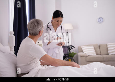 Infermiera asiatica a controllare il vecchio uomo. Bella donna asiatica in camice bianco con i suoi capelli in elegante bun verifica su paziente anziano della sua utilizzando gli appunti Foto Stock