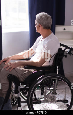 Triste uomo invecchiato in wheechair guardando fuori della finestra. Senior disabilitato l'uomo con i capelli grigi seduto in camera nella sua sedia a rotelle nella parte anteriore della finestra con marina cur Foto Stock