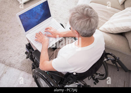 Uomo in sedia a rotelle digitando su laptop. Vista dall'alto di anziani disabili uomo con i capelli grigi utilizzando il computer mentre è seduto nella sedia a rotelle. Foto Stock