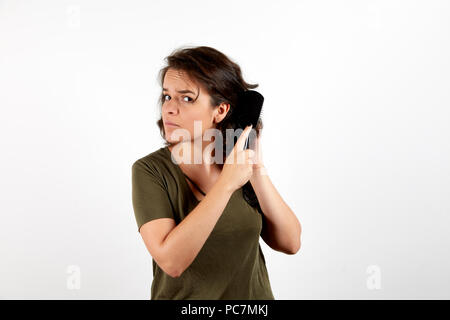 Bella giovane femmina conciata in casual MAGLIETTA SCURA wincing nel dolore durante la pettinatura scuro capelli aggrovigliati in piedi su bianco di sfondo per studio e alla ricerca di una Foto Stock