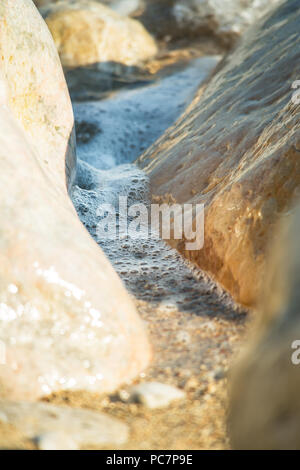 Una chiusura di bolle di una piccola onda / movimenti di marea lavaggio fino tra 2 roccia liscia su una spiaggia. Foto Stock