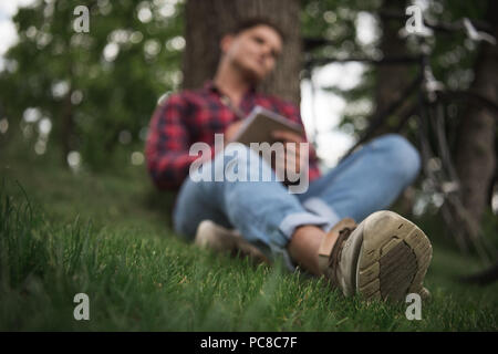 Giovane uomo prendendo appunti in notebook durante il riposo in posizione di stazionamento al giorno d'estate. Messa a fuoco selettiva su sneaker Foto Stock