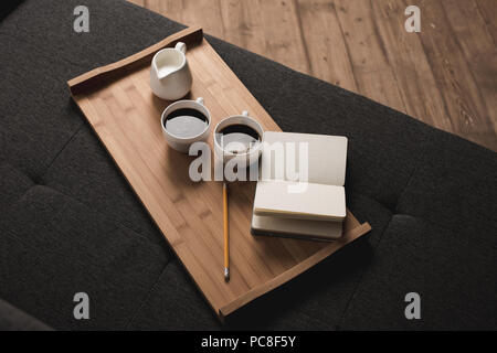 Due tazze di caffè, brocca del latte e notebook con matita sul vassoio in legno sul divano Foto Stock