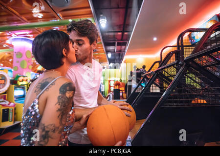 Paio di baciare ogni altro in piedi in un salotto di gioco tenendo basketballs. L uomo e la donna nella romantica atmosfera di un gioco arcade divertirsi giocando i giochi Foto Stock